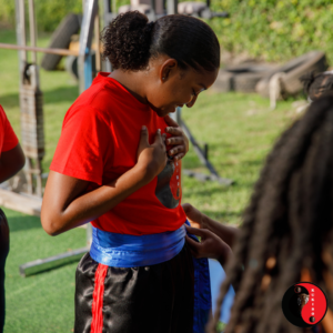 KUKIBO Student Lianne Receives Assistance With The Tying Of Her Sash On Her Person